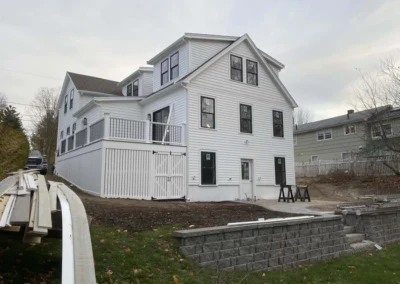 A large white house with black window frames and a spacious deck, surrounded by a yard with construction materials.