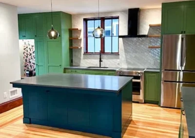 Modern kitchen with green cabinetry, blue island, stainless steel appliances, and wooden flooring.