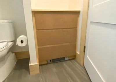 Bathroom with wooden cabinet blocking the paper roll and vent, next to a toilet and closed door.