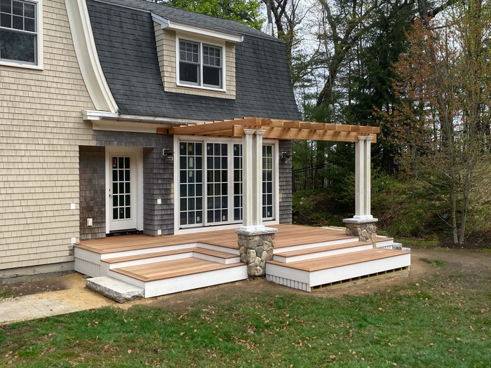 Custom carpentry deck with pergola attached to a house, featuring wooden steps and stone columns.