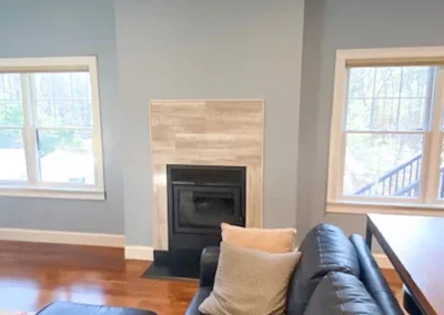 Living room with a black leather sofa, fireplace, and two windows with a view of trees.