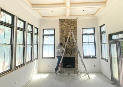 Worker on ladder constructing a stone fireplace in a bright room with large windows.