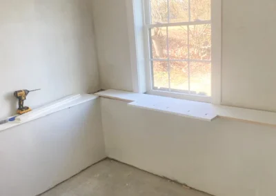 Drill and unfinished shelving under a window in a bright, white room.