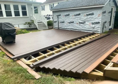 Wooden deck partially built, with planks stacked and framing exposed, next to a white house and garage under construc...