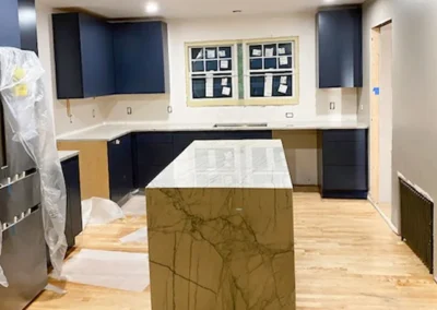 Modern kitchen with dark cabinets, marble island, and wood flooring. Plastic covers the fridge.