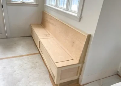 Wooden storage bench with hinged seats beneath windows in a sunlit room.