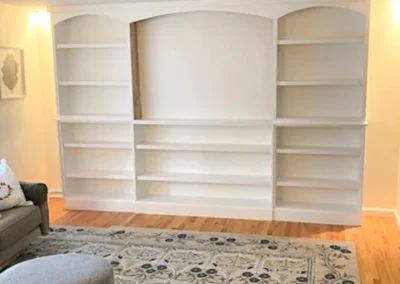 Empty white built-in bookshelves with arched tops on a light-colored wall, next to a sofa and a patterned rug.