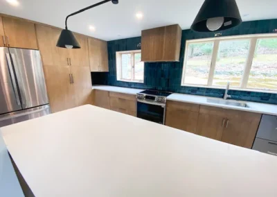 Modern kitchen with wood cabinets, white counters, teal backsplash, stainless appliances, and black pendant lights.