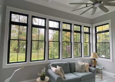 A cozy living room with a large sofa, pillows, and a lamp, featuring multiple tall windows offering a view of lush greenery outside.