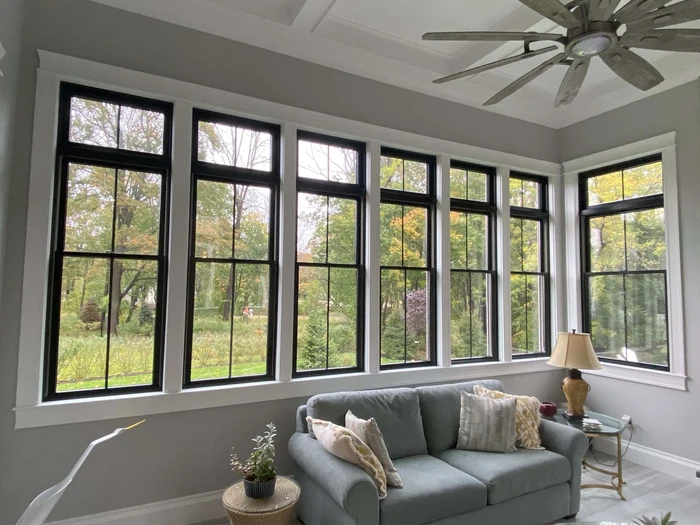 A cozy living room with a large sofa, pillows, and a lamp, featuring multiple tall windows offering a view of lush greenery outside.