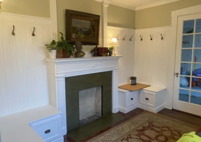 Cozy room with custom carpentry featuring a white mantle and built-in benches surrounding a fireplace, with decorative hooks on the beadboard walls.