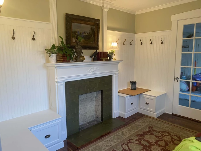 Cozy room with custom carpentry featuring a white mantle and built-in benches surrounding a fireplace, with decorative hooks on the beadboard walls.