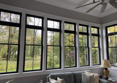 A sunroom with a series of large, black-framed windows offering a view of a green, tree-filled landscape. Inside, there is a comfortable gray couch with decorative pillows and a lamp.