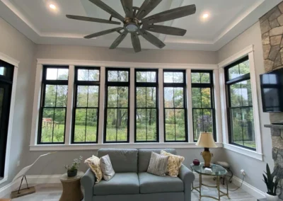 A cozy living room with large windows, a sofa, and a ceiling fan, offering a view of a lush green outdoor area.