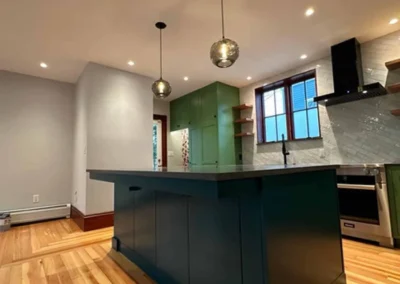 Modern kitchen interior with wooden flooring, dark green island, and two pendant lights.