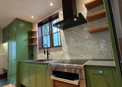 A modern kitchen featuring green cabinets, silver appliances, and wooden shelving against a tiled backsplash.
