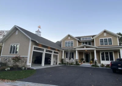 A large, modern two-story house with beige siding and white trim. The home features a spacious front porch with decorative pillars, multiple large windows, and a three-car garage. A decorative weathervane sits atop a cupola on the roof. A black truck is parked in the driveway and a ladder is leaned against the porch.